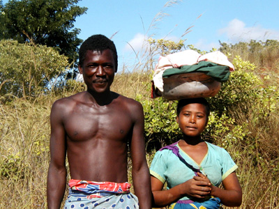 Two people standing beside vegetation, one carrying a bundle on their head